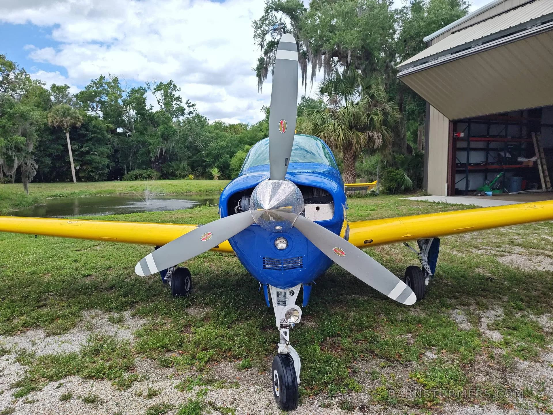 A blue and yellow single-engine aircraft is viewed from the front, showing its propeller and nose gear, parked in front of a hangar and pond.