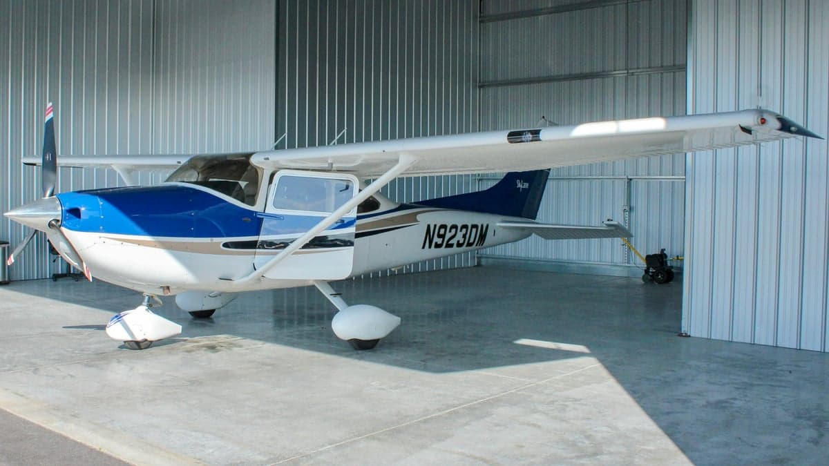 A white and blue single-engine propeller aircraft, N923DM, parked inside a metal hangar, viewed from the front-left.