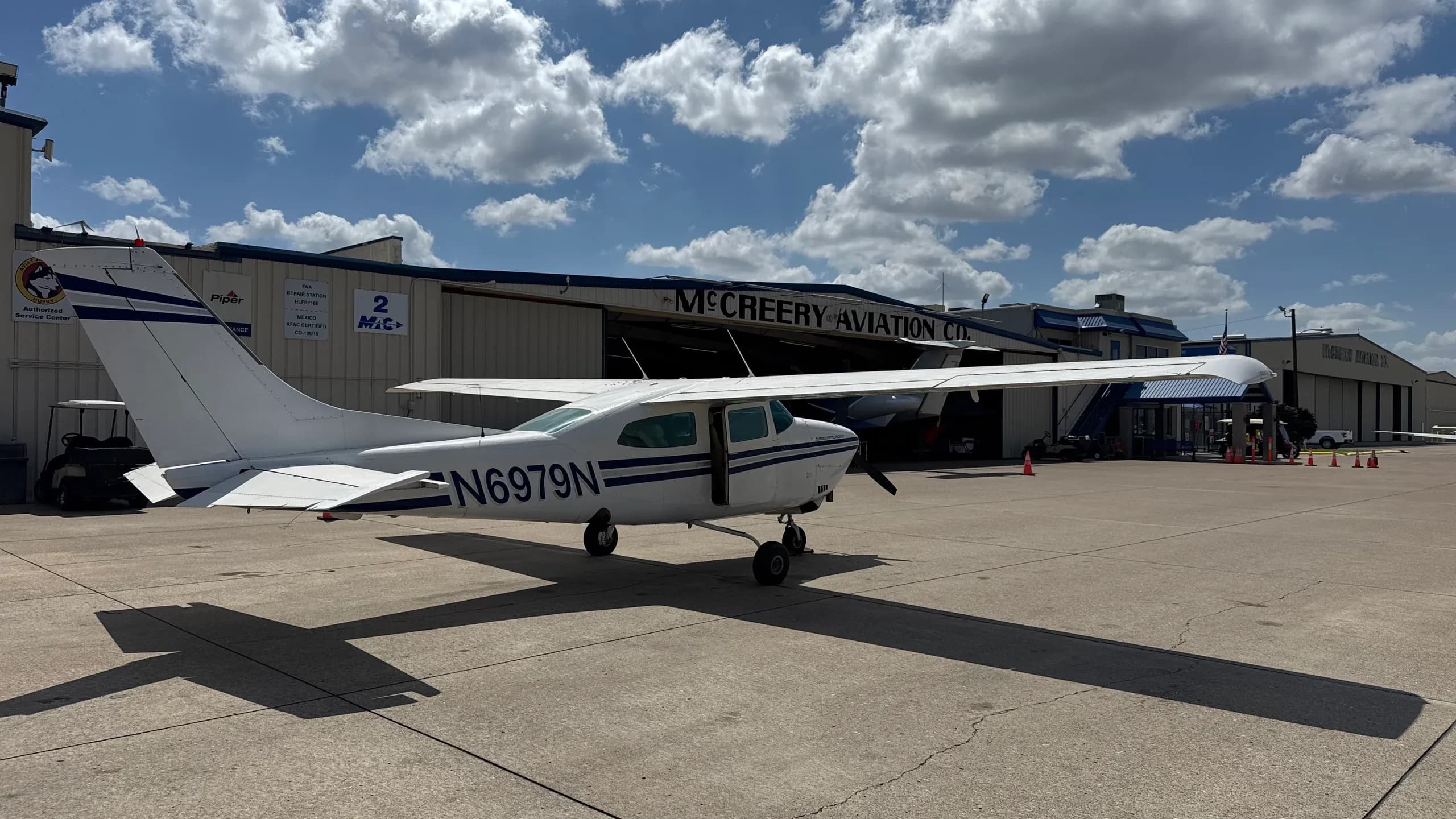A rear-side view of a white Cessna 182 with blue stripes, registration N6979N, parked on a tarmac in front of a hangar.
