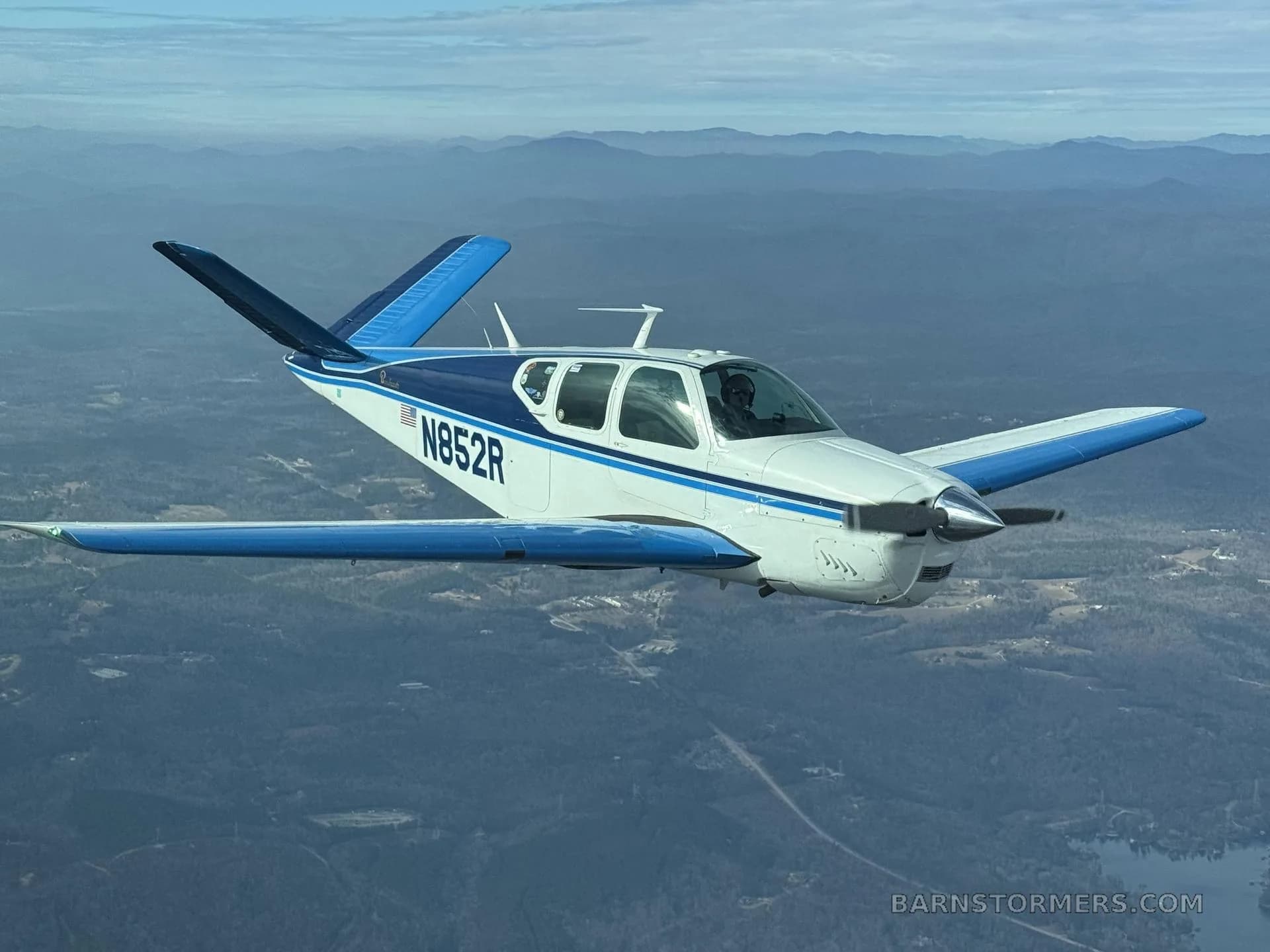 A white and blue Beechcraft Bonanza N852R flies over a mountainous, forested landscape under a blue sky.
