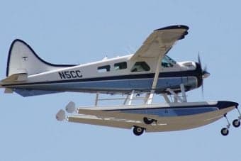 A white and silver float plane takes off from a lake with mountains in the background.