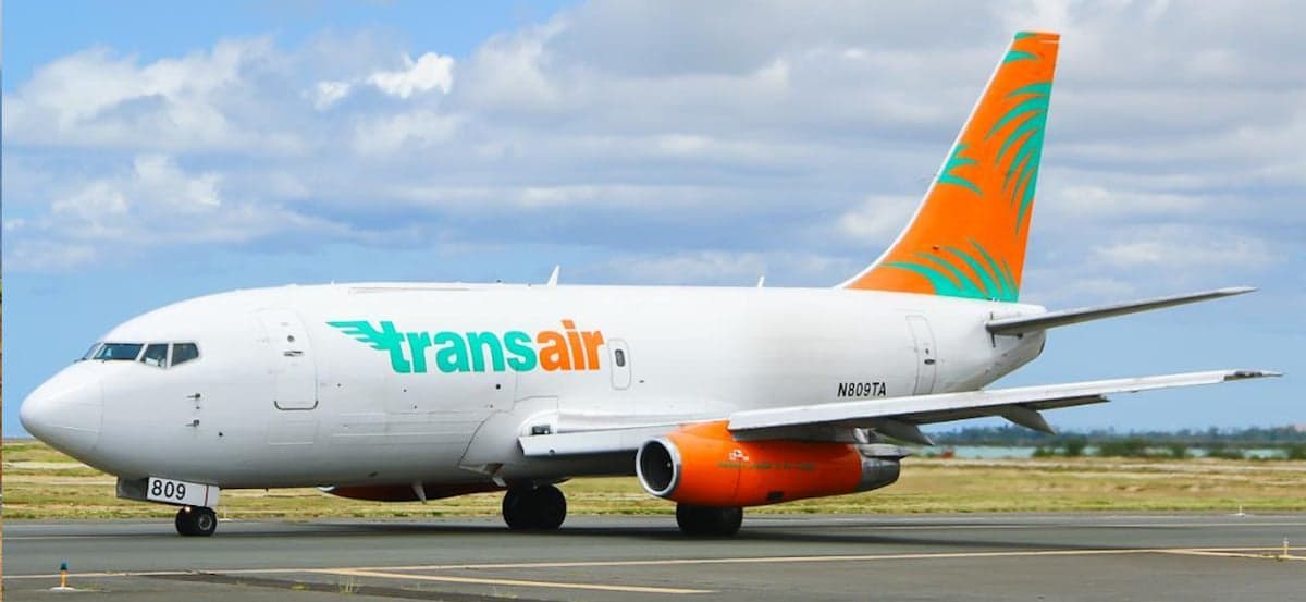 Side view of a white Boeing 737 aircraft with an orange and green stripe on the tail, parked on the tarmac.
