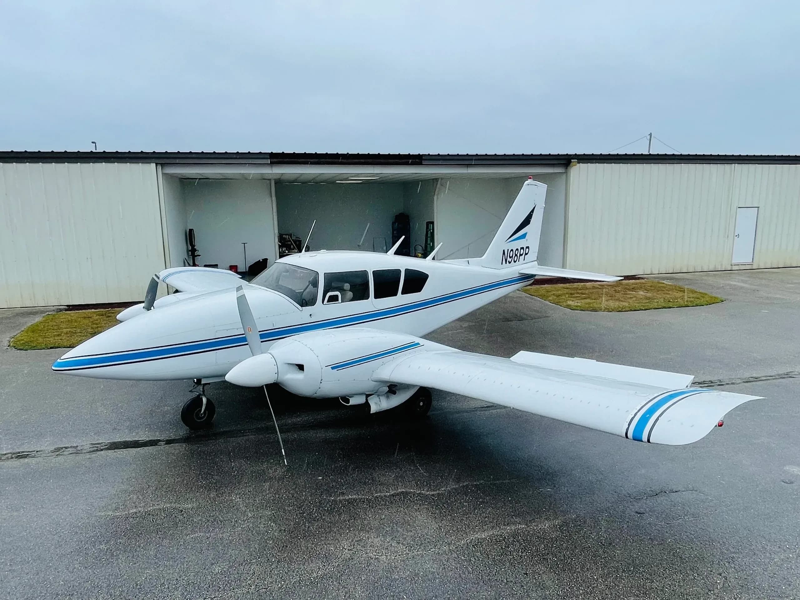 A white Piper Aztec aircraft with blue stripes parked on a wet tarmac in front of an open hangar.