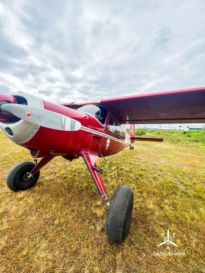 A low-angle view of the front-right side of a red and silver single-engine aircraft with large tires, parked on a grassy field.