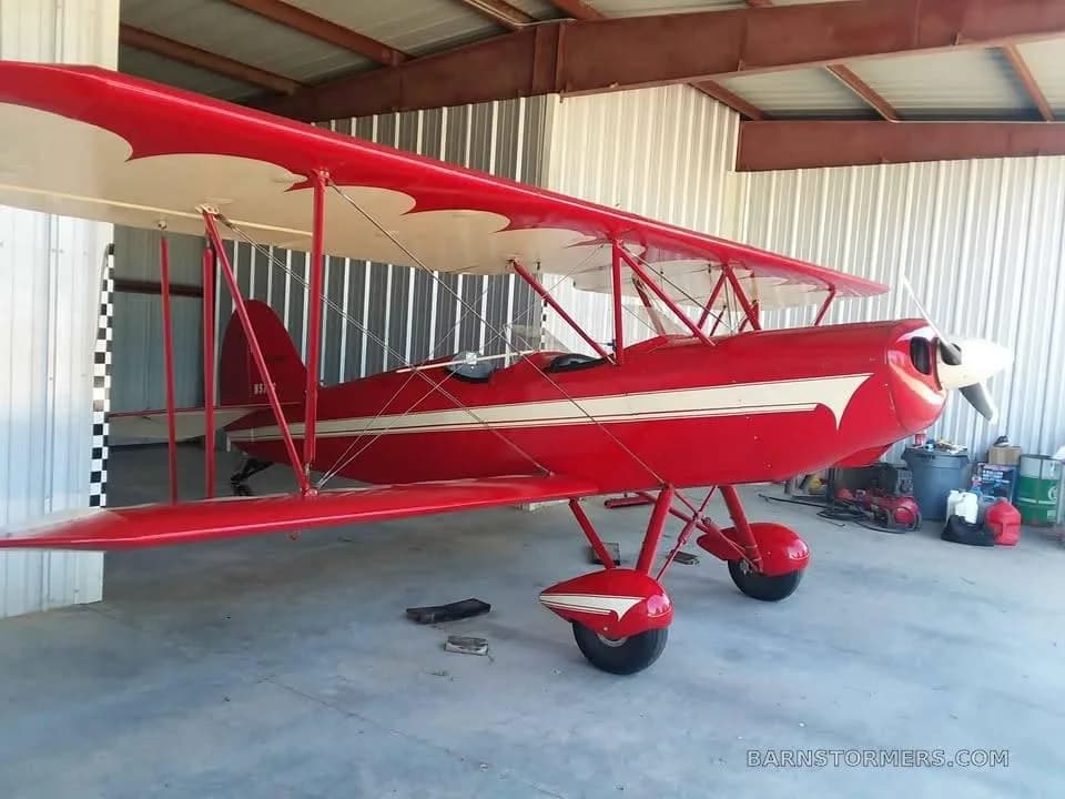 A red biplane with white stripes and wing accents, parked inside a metal hangar.