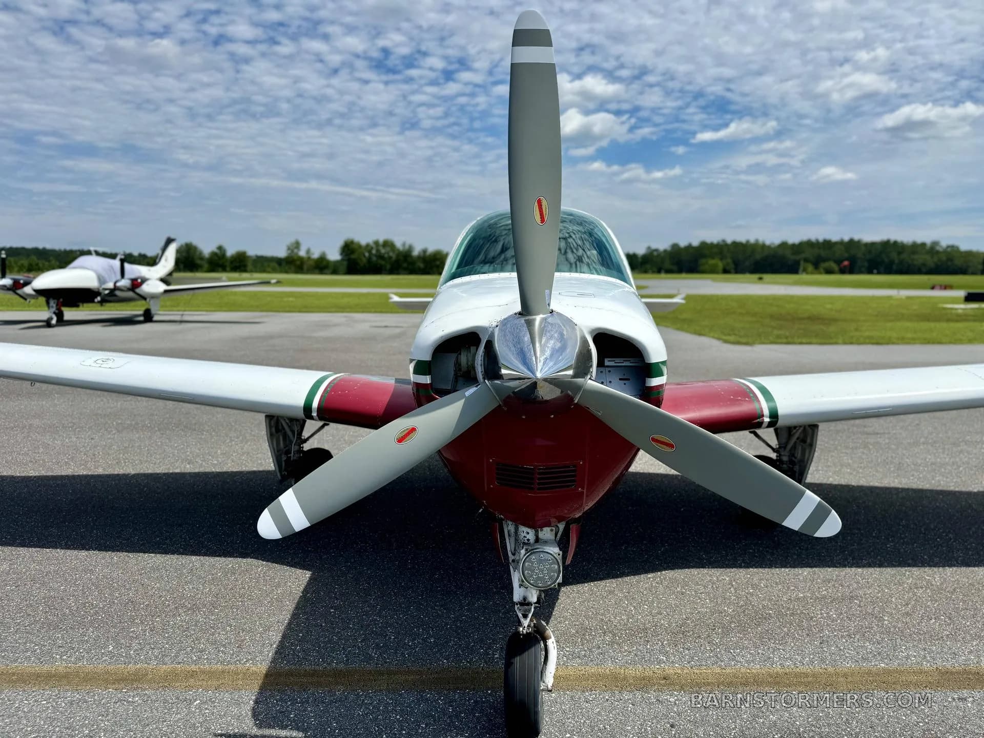 A close-up front view of the propeller and nose of a white and maroon single-engine aircraft on an airfield.