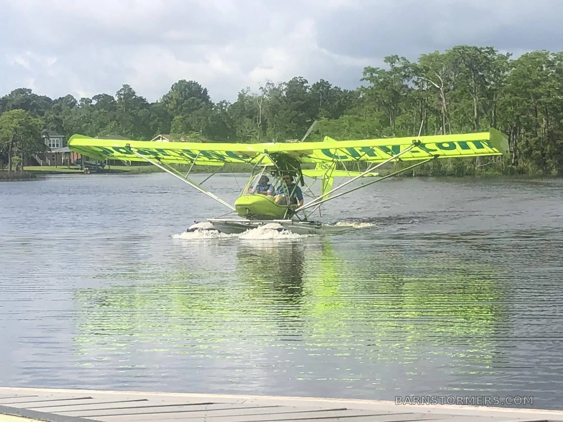 A bright green ultralight seaplane with two occupants creates a wake as it moves across the water, with trees in the background.