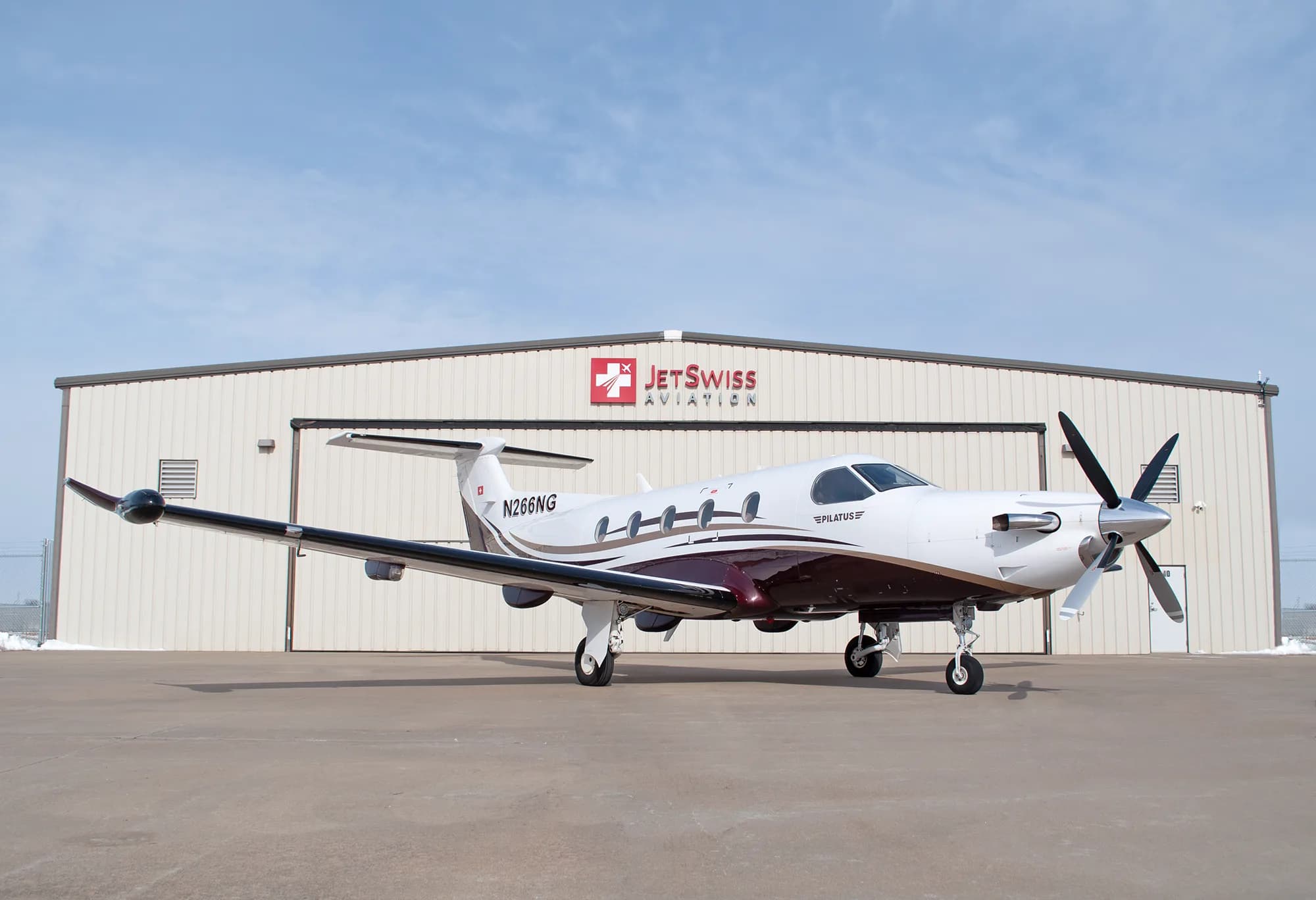 A Pilatus PC-12 NG aircraft with white and brown paint scheme, registration N266NG, parked on a tarmac in front of a hangar.