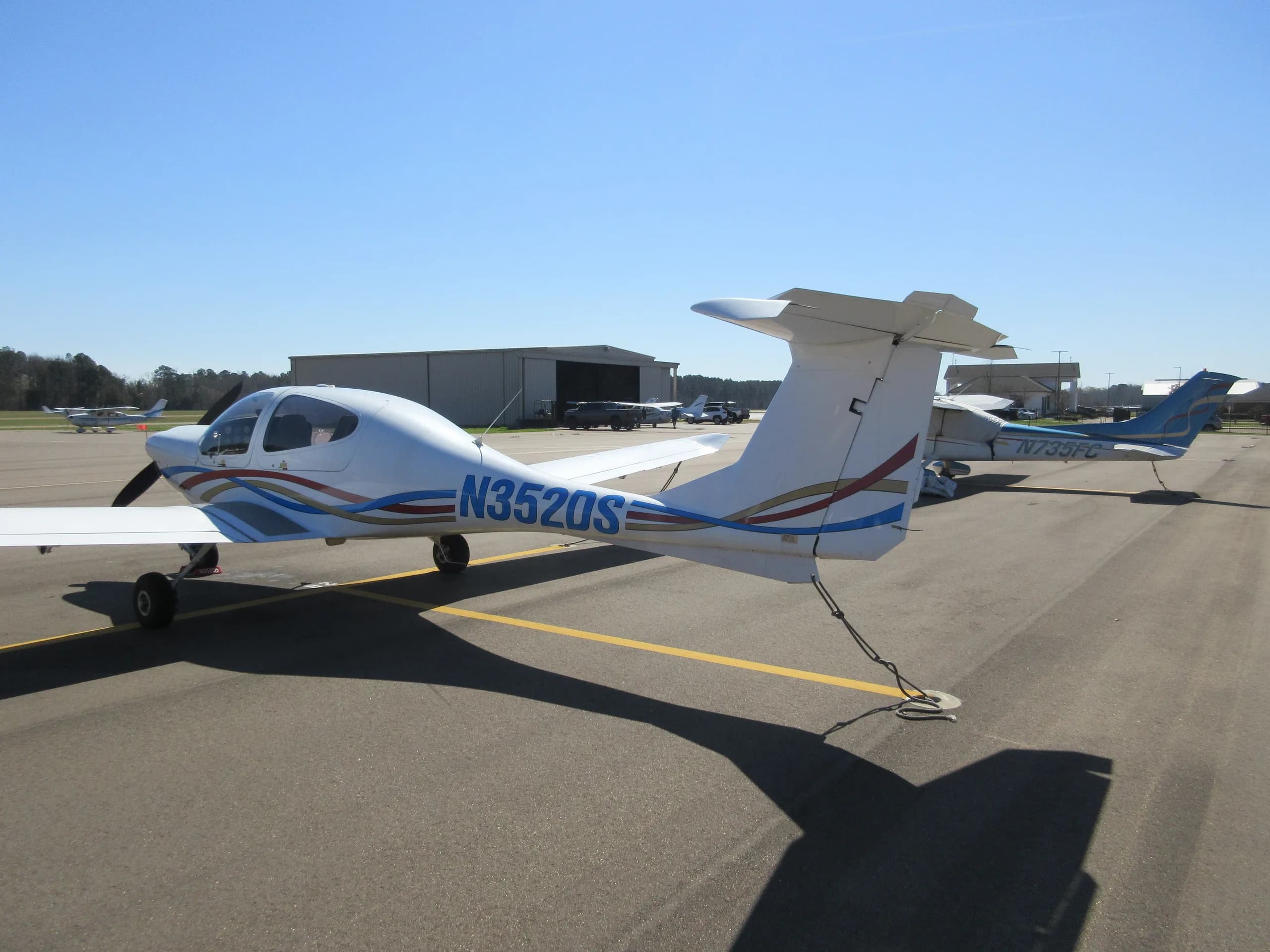 A white Diamond DA40 aircraft with blue, red, and gold stripes, registration N352DS, parked on an airport tarmac.