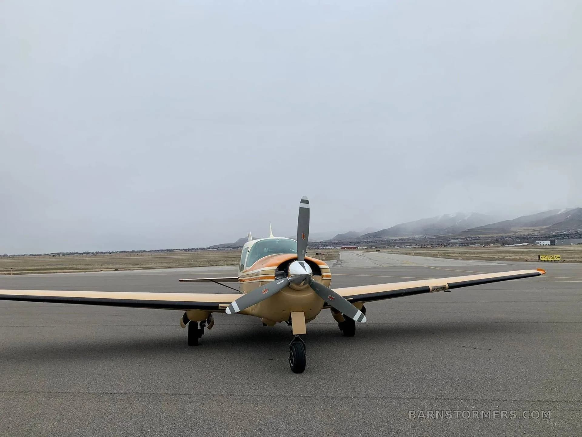 A beige and orange single-engine aircraft (N625RL) parked on a runway, viewed from the front, with mountains in the background under a cloudy sky.
