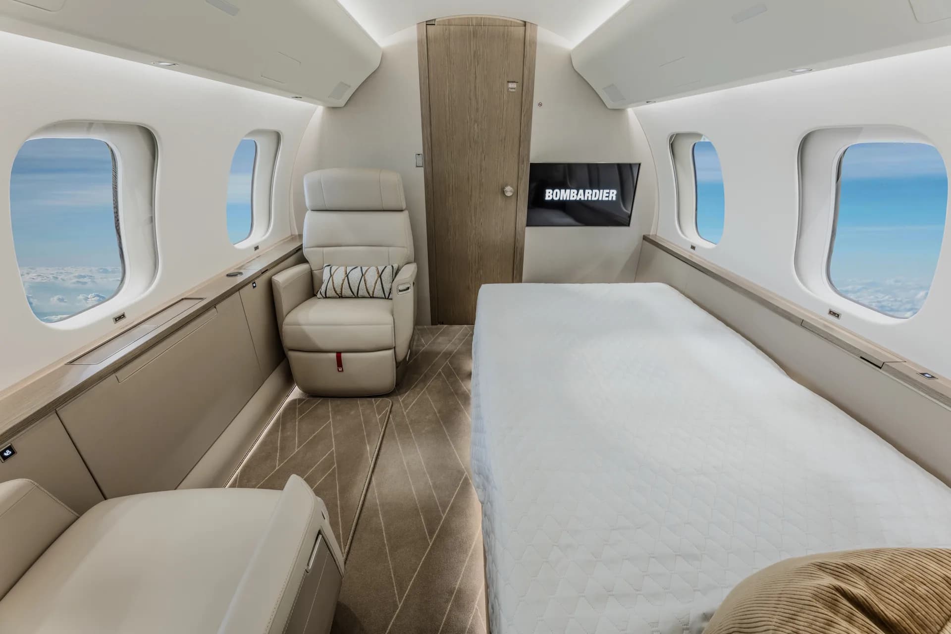 A private jet cabin featuring a bed on the right and a cream leather seat on the left, with a Bombardier screen.