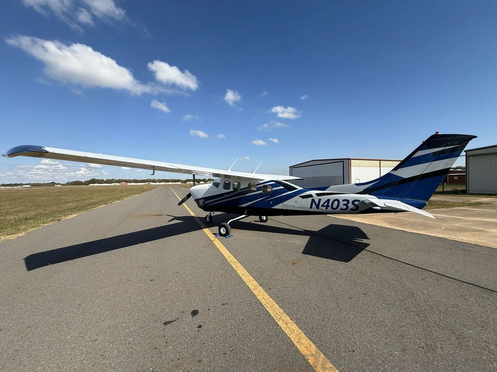 A white and blue Cessna 210 Centurion aircraft (N403S) parked on an airfield with a blue sky and clouds.