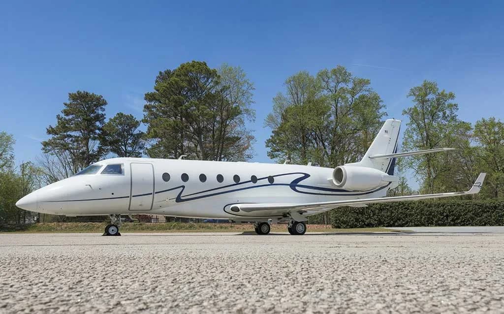 A white business jet with blue wavy stripes on the fuselage, parked on an asphalt surface with trees in the background.