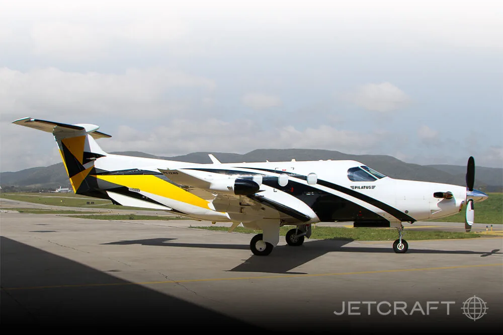 A white, black, and yellow Pilatus PC-12 aircraft parked on an airfield with mountains in the background.