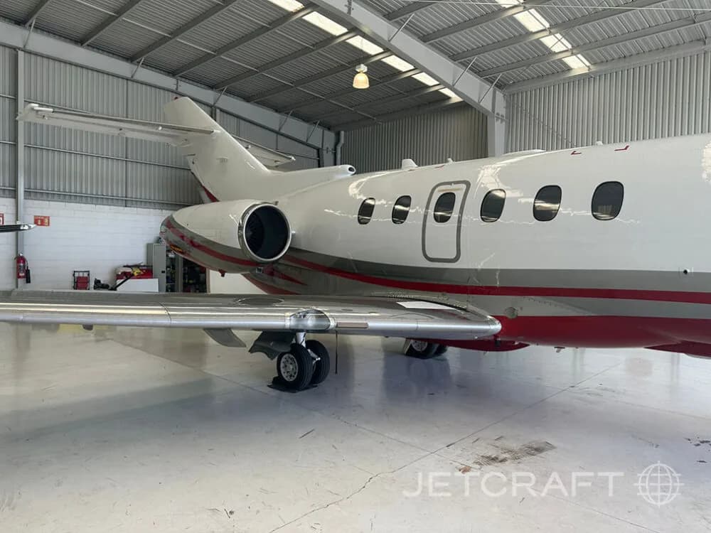 A side view of a white and red business jet with its wing and landing gear visible, parked in a hangar.