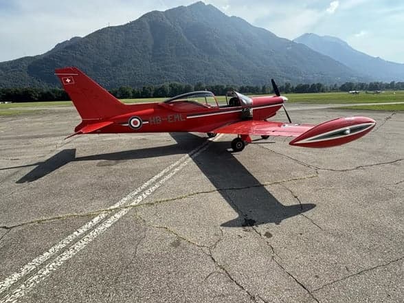 A red aircraft, HB-EML, parked on the tarmac with mountains in the background.
