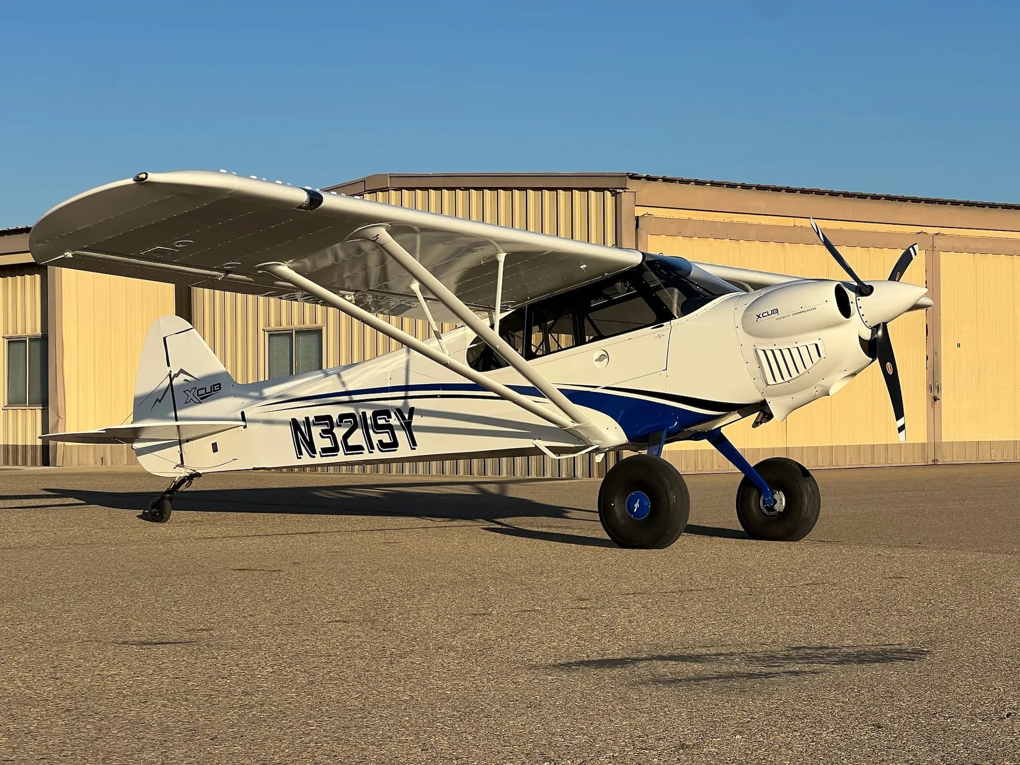 A white X-Cub aircraft with blue stripes and registration N321SY parked on an airfield with a building in the background.