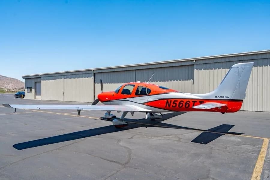 Side view of a red and silver Cirrus SR22T aircraft (N566TT) parked on an airfield with hangars in the background.