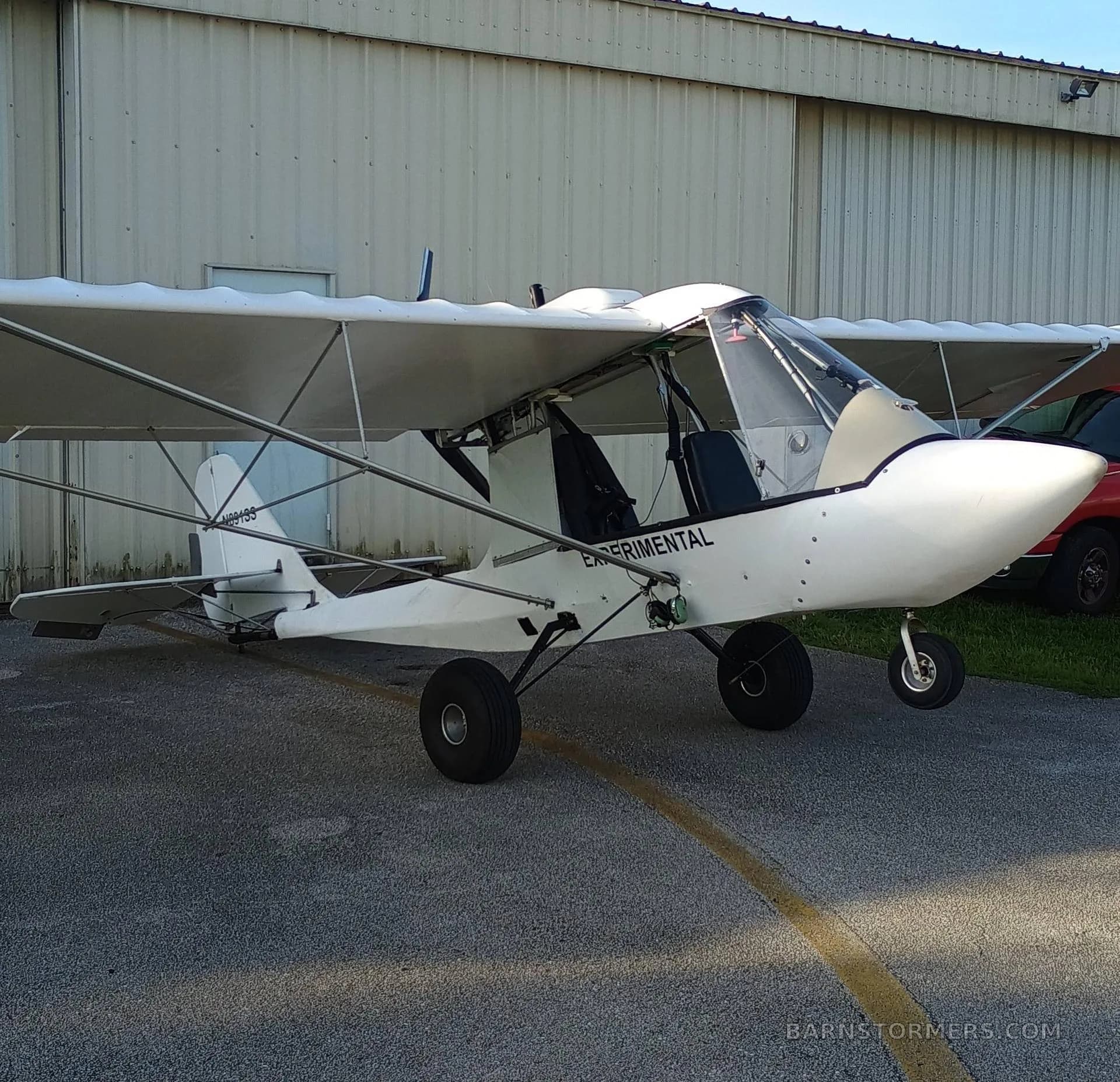 A white experimental ultralight aircraft with a high wing is parked on asphalt in front of a metal hangar, seen from the front-right side.