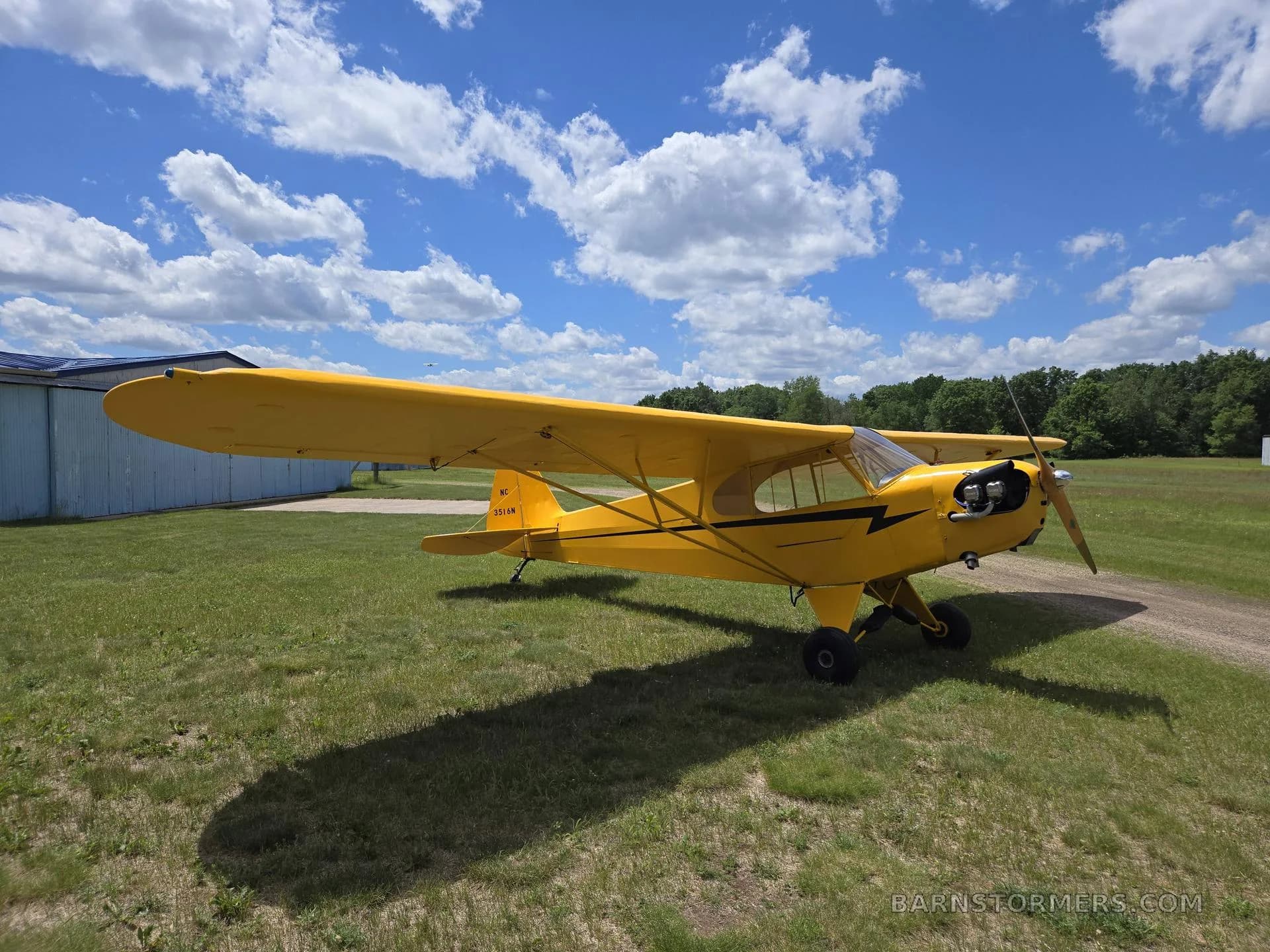 A yellow Piper Cub-style aircraft with black lightning bolt accents is parked on a grassy field under a blue sky.