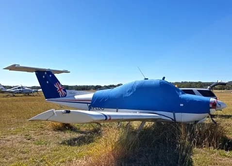 A white Beechcraft Skipper 77 with a blue cockpit cover and Australian flag tail, parked on a grassy field under a clear blue sky.