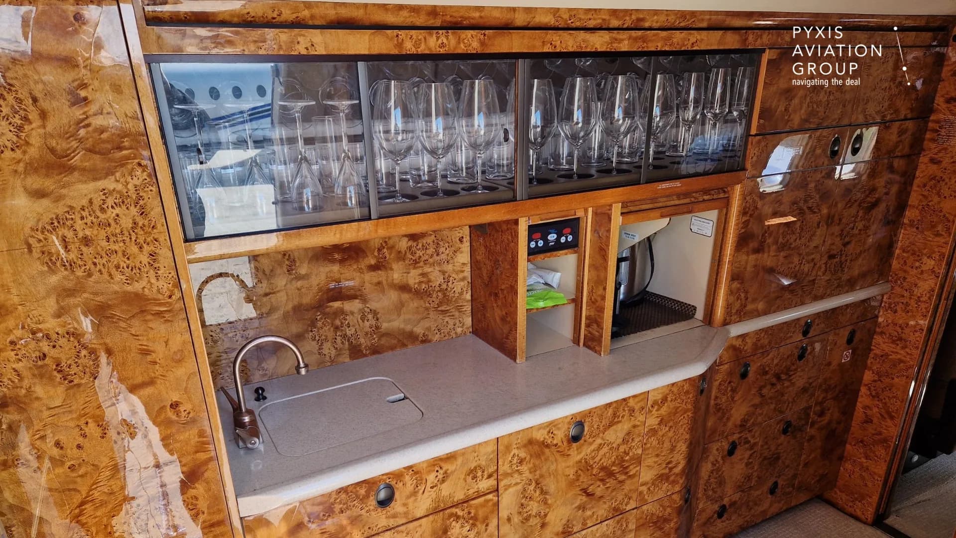 A close-up of the aircraft's galley counter with a sink, coffee maker, and glass storage cabinets filled with glassware.