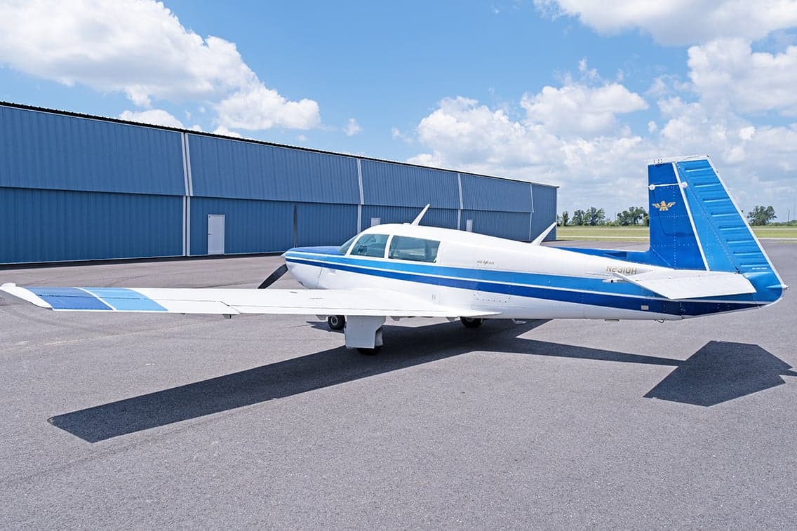 Side view of a white and blue Mooney M20F aircraft parked on the tarmac in front of a hangar.