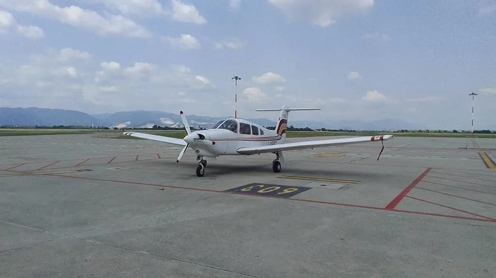 A white Piper PA-28 aircraft with red and gold stripes parked on an airfield with mountains in the background.
