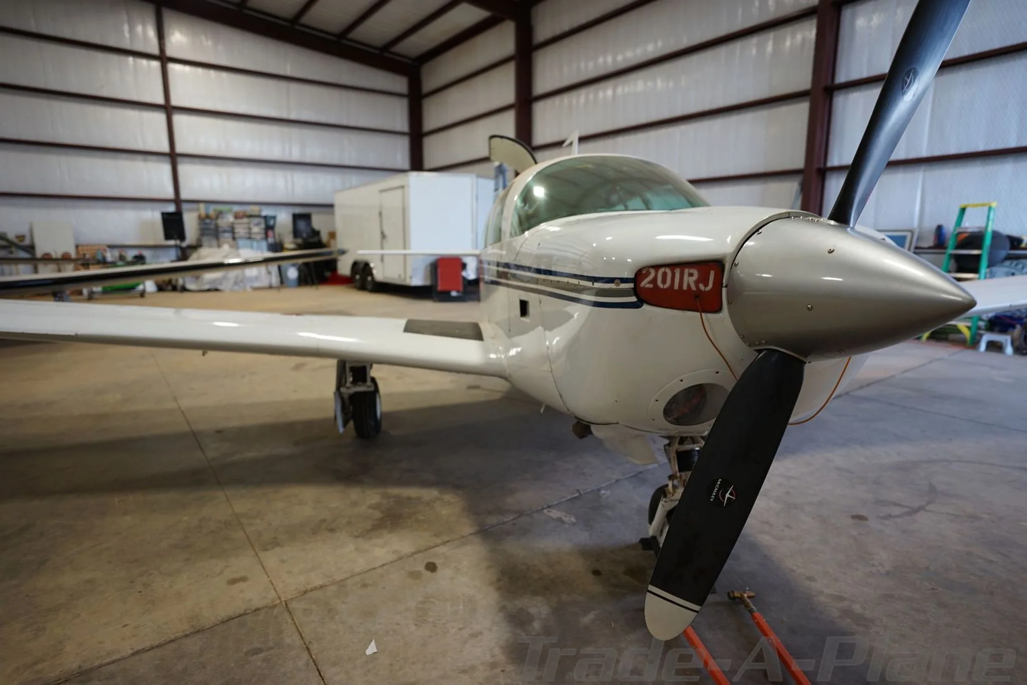 A front view of the white single-engine aircraft N201RJ with a silver spinner and black propeller, parked in a hangar.