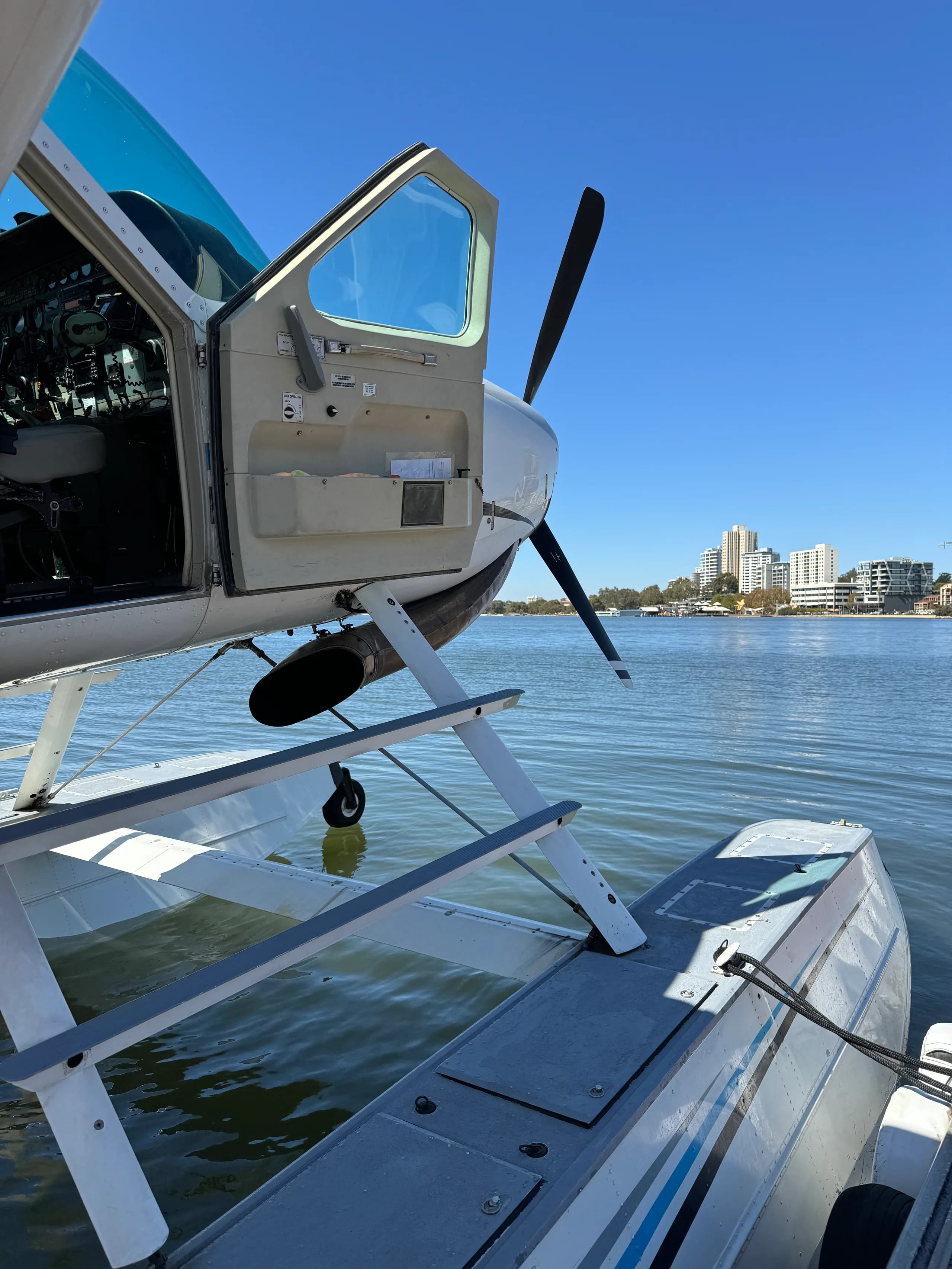 A white seaplane with an open door is docked in calm water, with city buildings visible across the water under a clear blue sky.