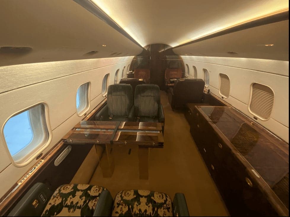 A view down the aisle of the aircraft cabin, showing dark wood cabinetry, a fold-out table, and patterned seats.