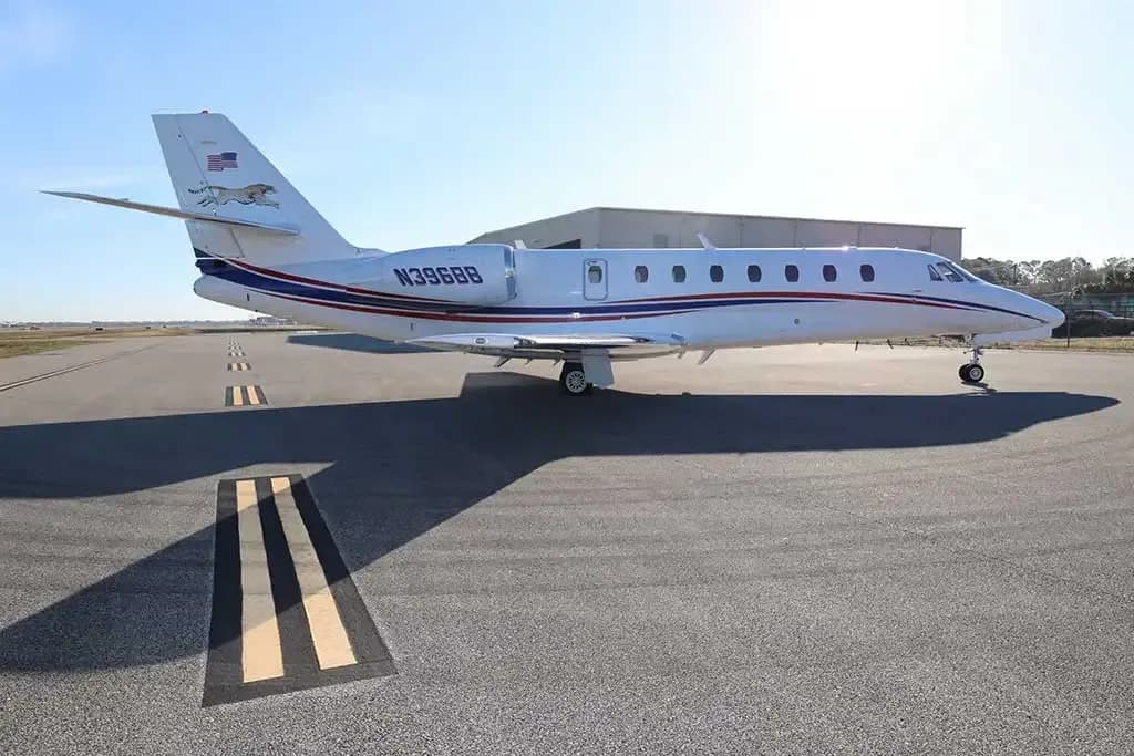 A side view of the white Cessna Citation business jet N396BB with red and blue stripes, parked on an airfield with a building in the background.