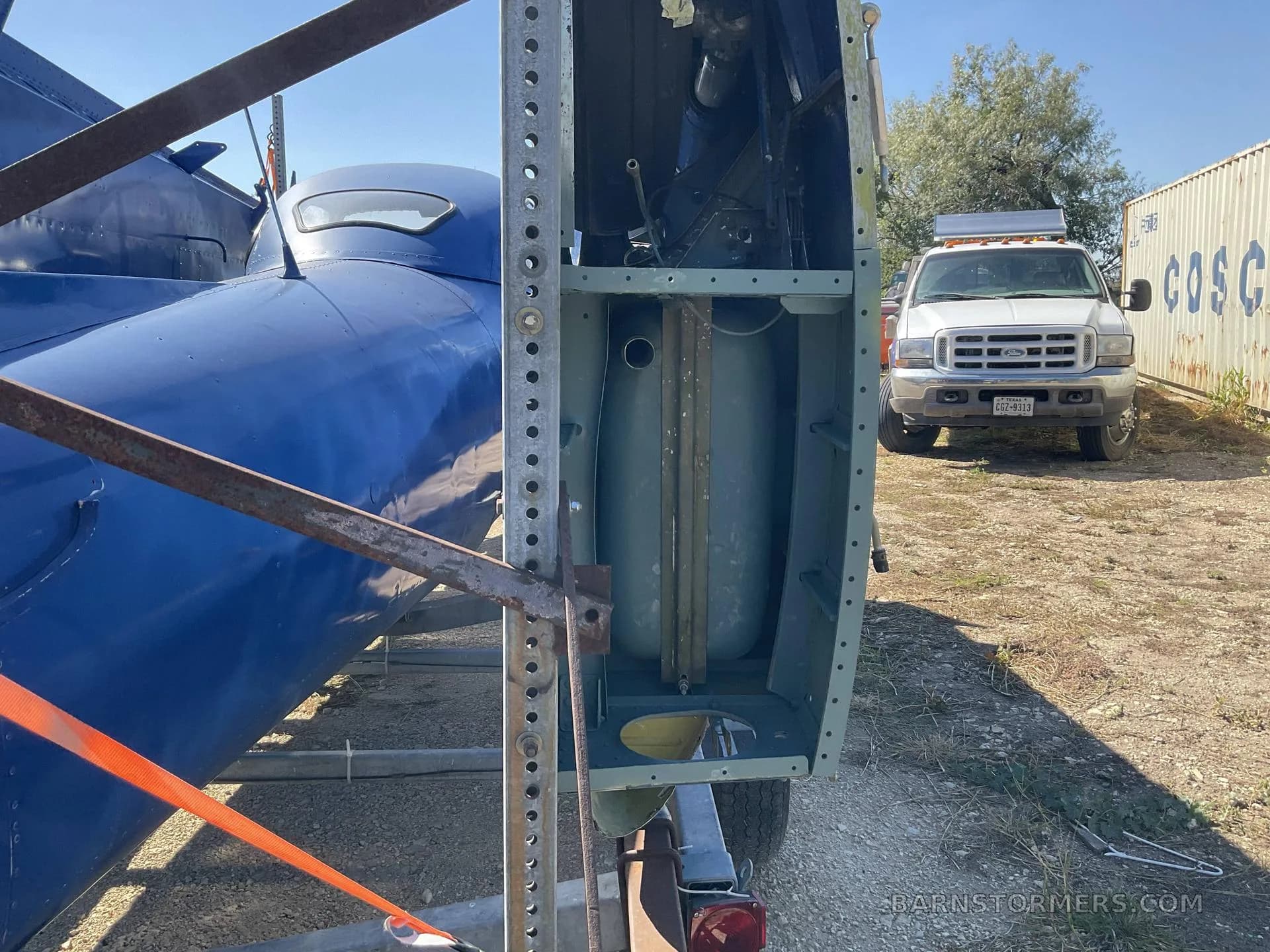 A close-up view of the rear section of a blue aircraft fuselage on a trailer, revealing internal structure and a fuel tank, with a white pickup truck in the background.