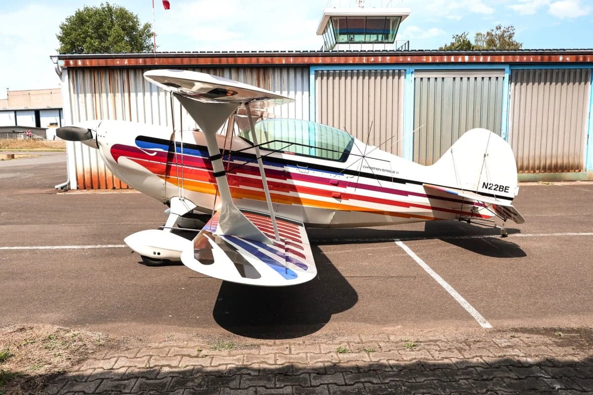 Side view of the Christen Eagle II biplane with colorful stripes, parked on the tarmac near a building.