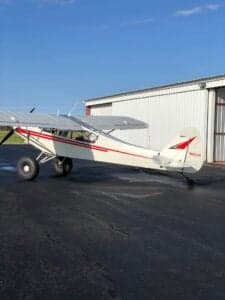 A white and red Piper Cub sits on the tarmac in front of a hangar.