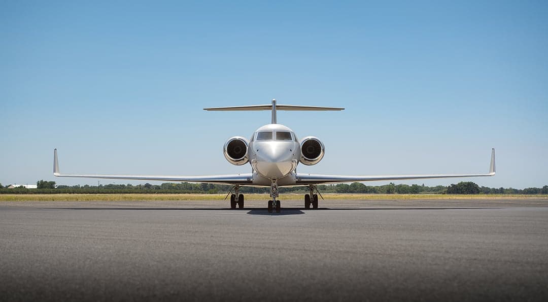 A head-on view of a white Gulfstream G550 business jet with two engines, parked on an airfield under a clear sky.