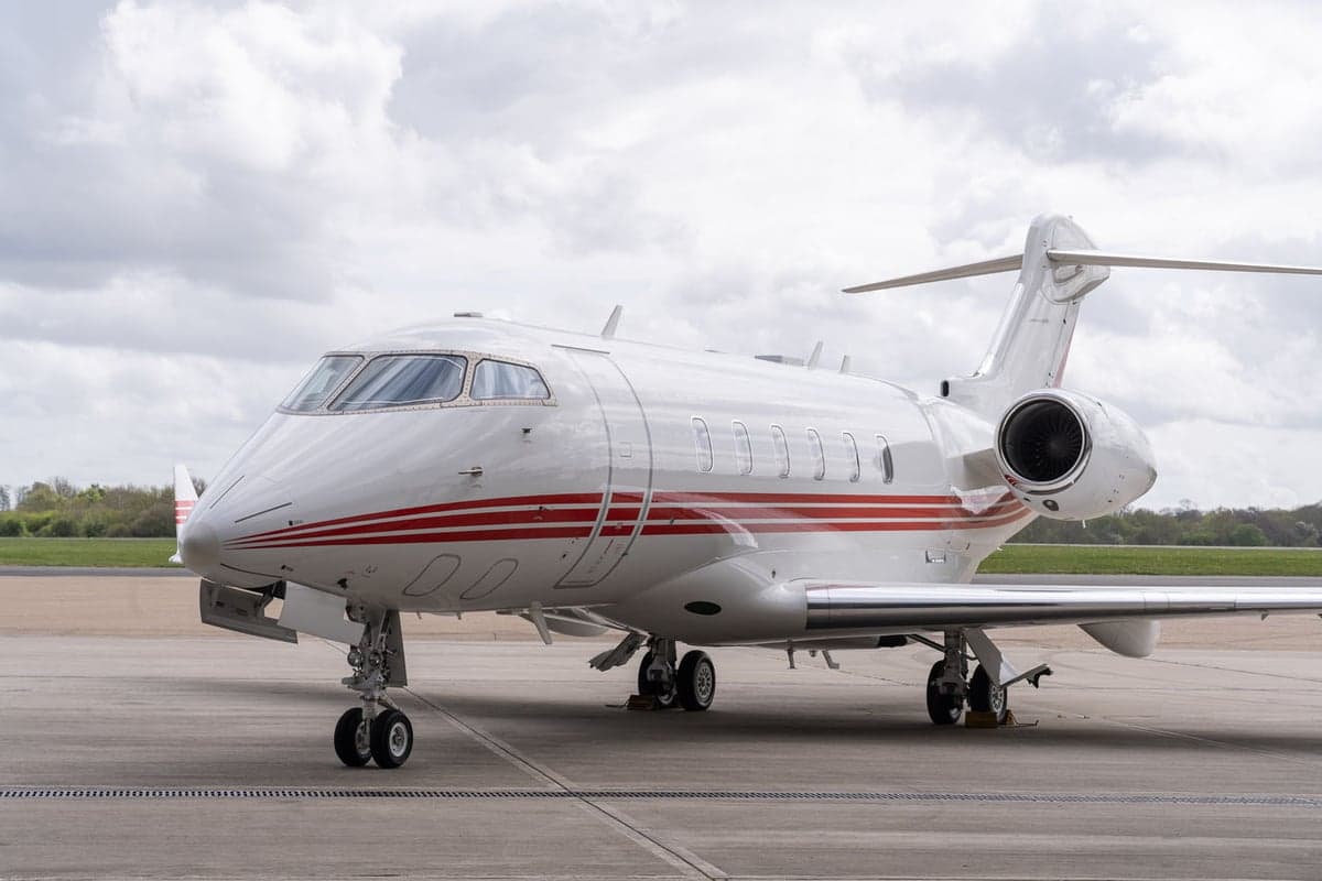 A white business jet with red stripes parked on an airfield under a cloudy sky, viewed from the front-right.