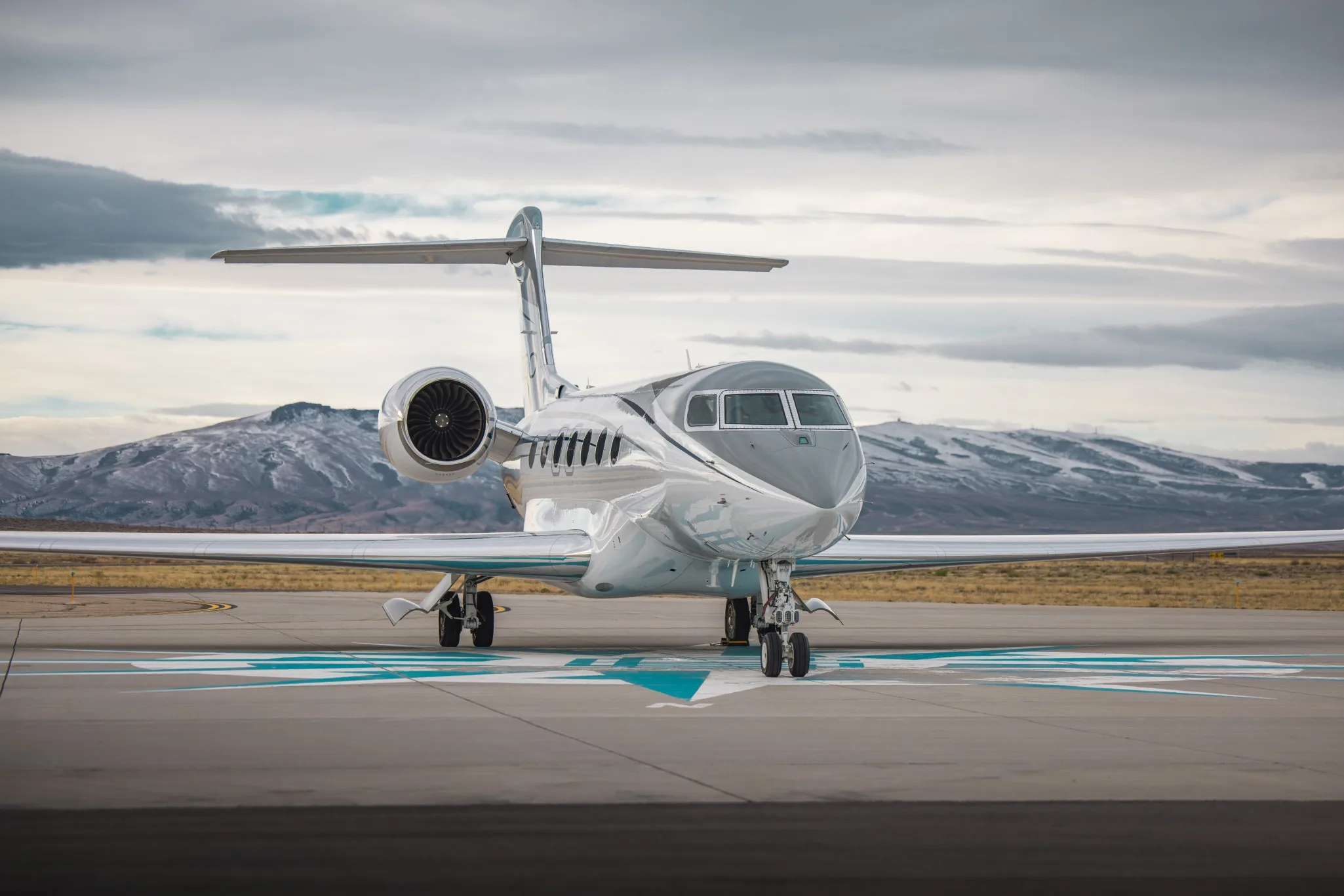 A silver Gulfstream business jet with a grey nose parked on an tarmac with blue markings and snowy mountains in the background.