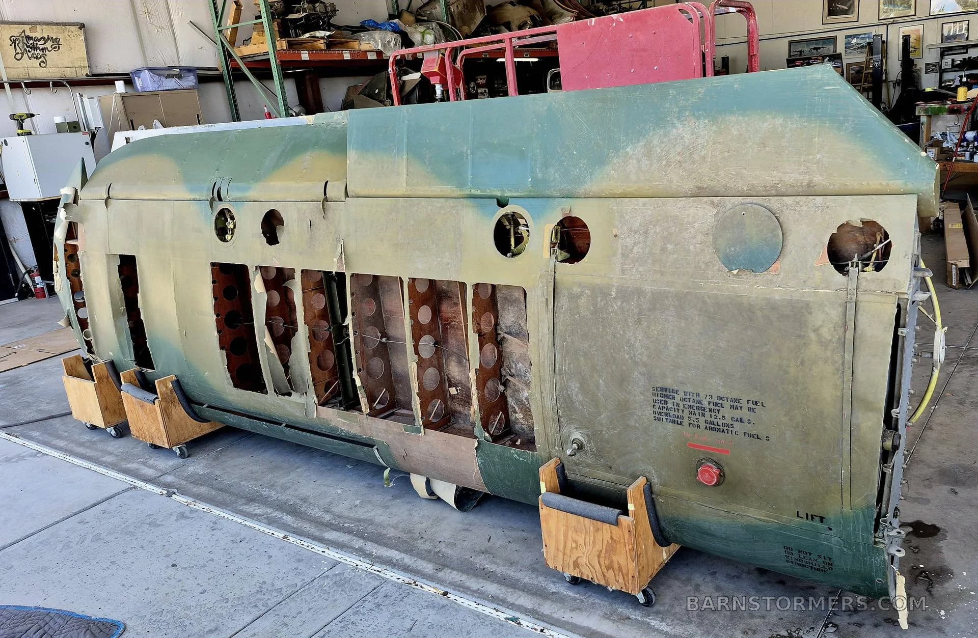 A large, olive-green fuselage section with exposed internal structure and stenciled text, resting on wooden supports in a hangar.