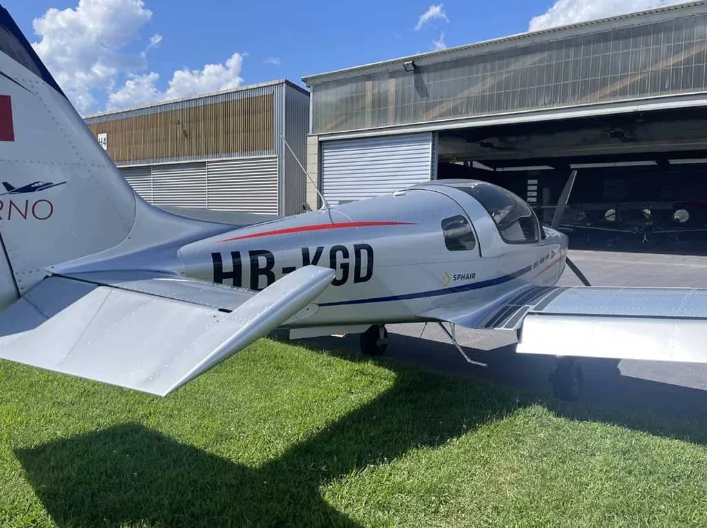 A close-up view of the silver tail and rear fuselage of the Sonaca 200 aircraft (HB-KGD) parked near hangars.