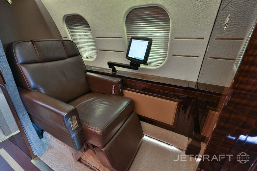A brown leather seat next to a window, with a small monitor on a dark wood side table in the business jet cabin.