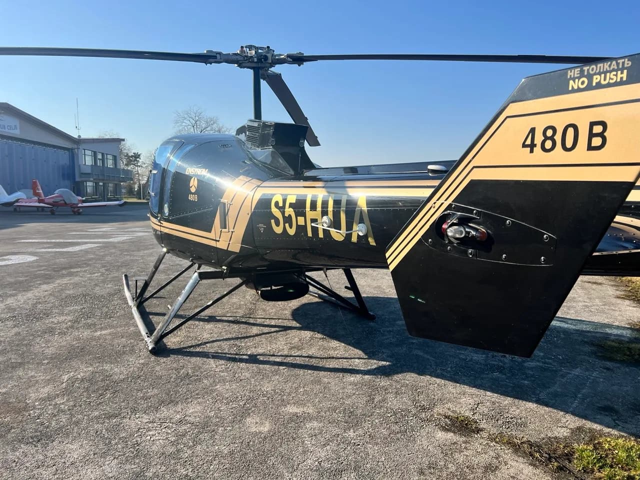 A rear-side view of the black and gold Enstrom 480B helicopter (S5-HUA) parked near a hangar.