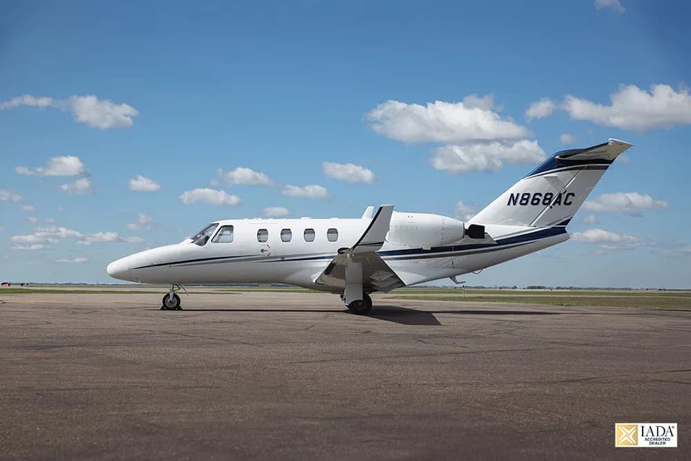 A white Cessna Citation CJ1+ (N868AC) with blue and grey stripes parked on an airfield under a partly cloudy sky.