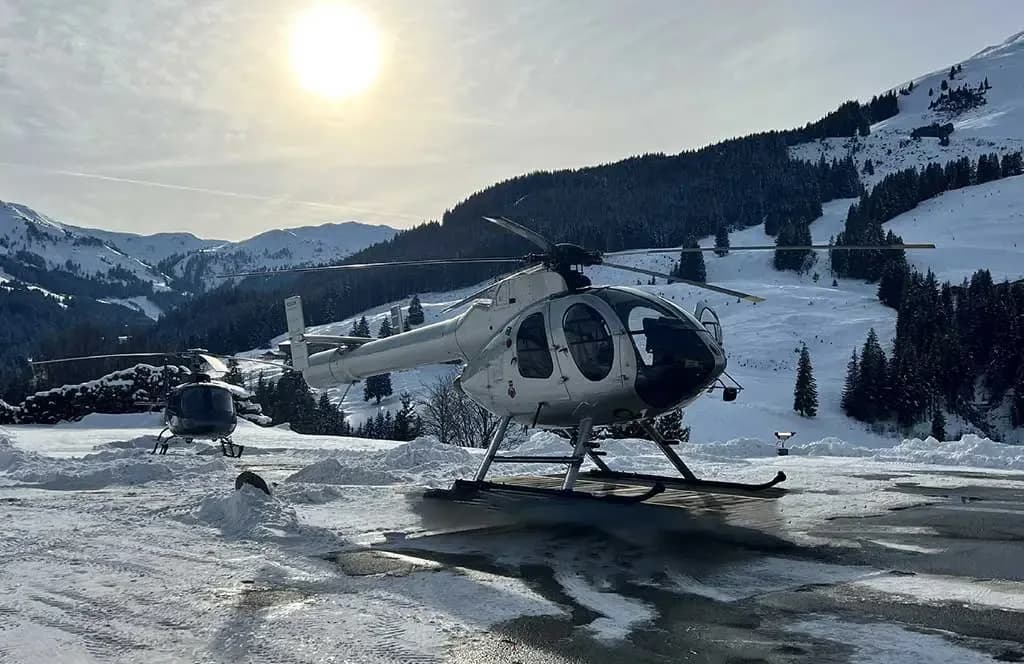 Two silver helicopters are parked on a snow-dusted tarmac with snowy mountains and a bright sun in the background.