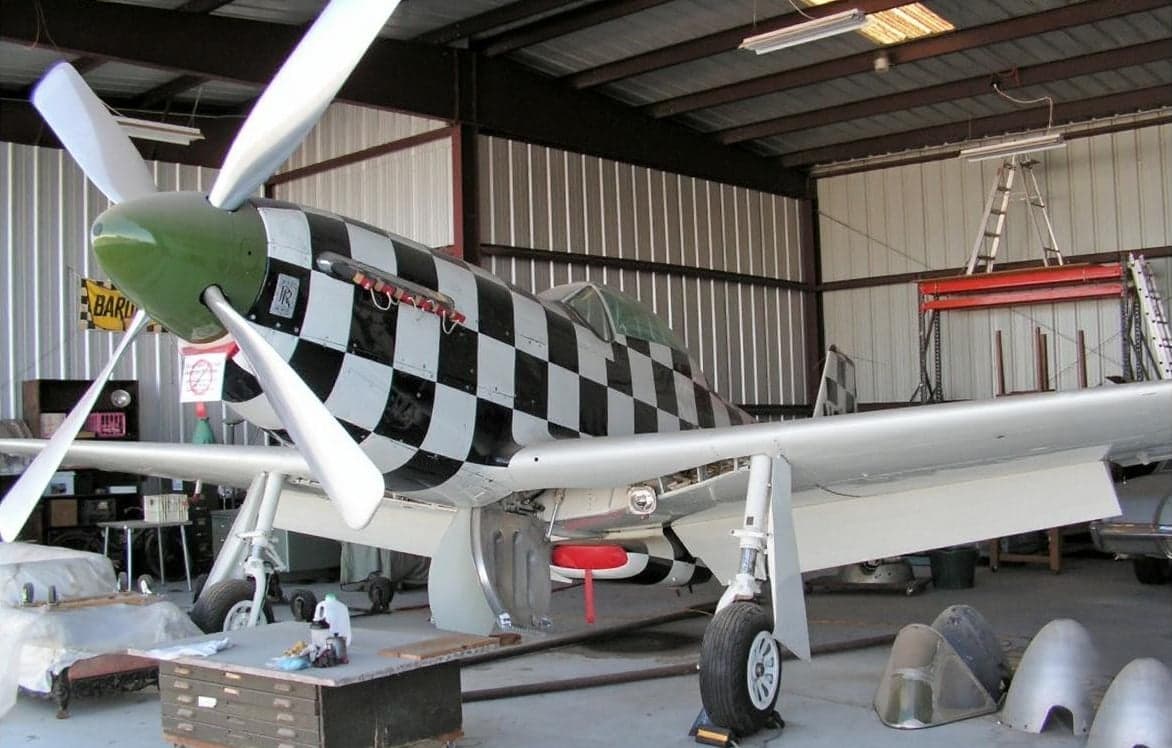 A P-51 Mustang with a black and white checkered paint scheme is parked in a hangar.