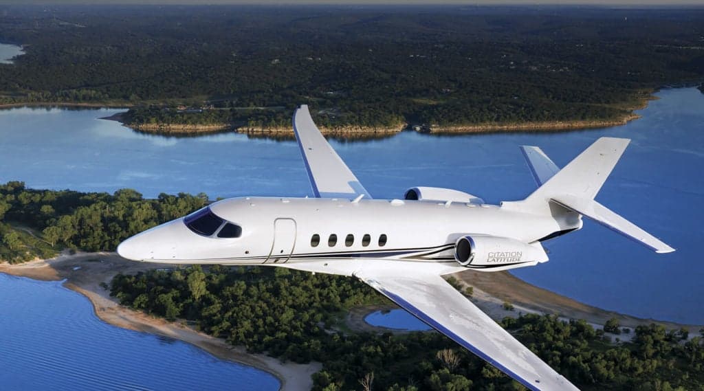 A Citation Latitude jet flying over a lake and forest landscape.