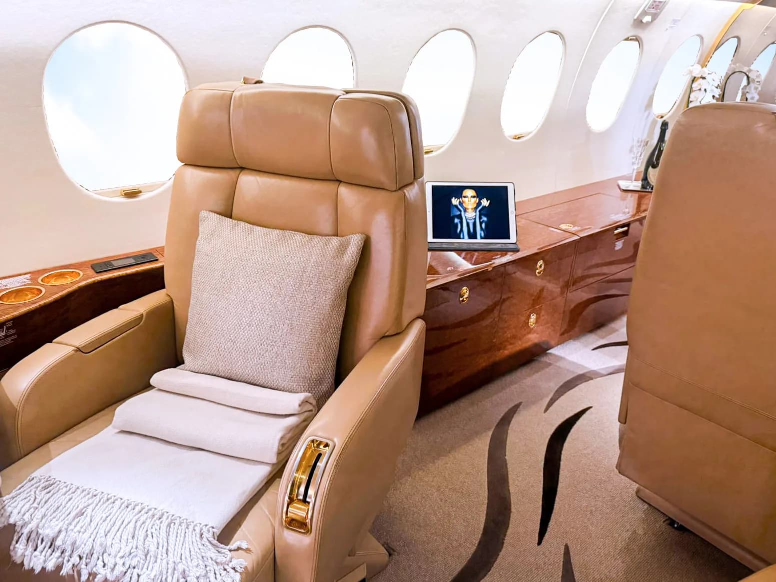 A tan leather seat with a throw blanket and pillow is seen in a private jet cabin, with a laptop on a wooden console.