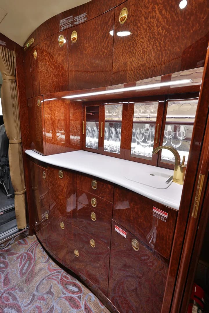 A luxurious aircraft galley features dark wood cabinetry, a white countertop with a sink, and glass-fronted cabinets displaying glassware.