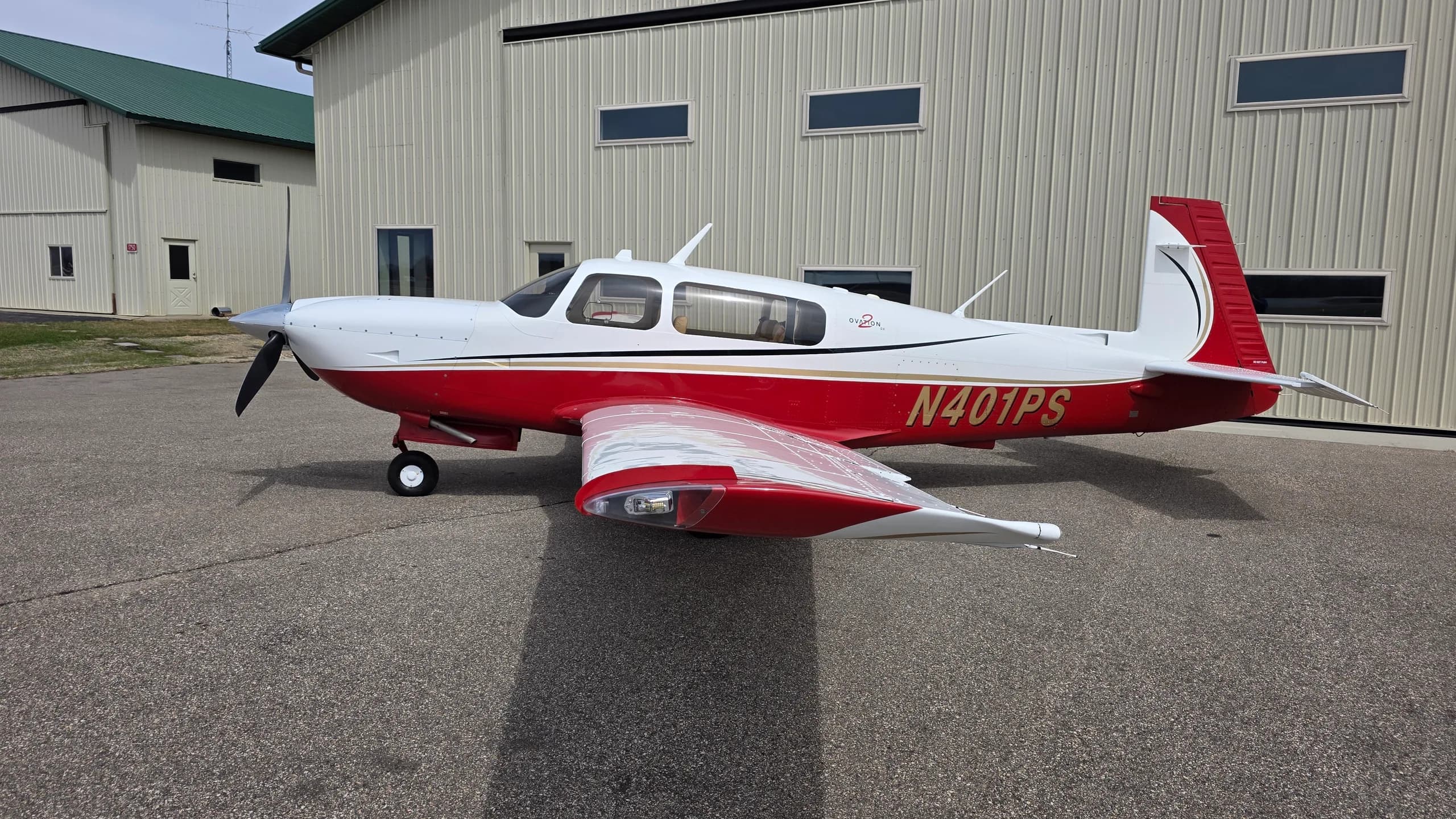 A full side view of a red and white Mooney aircraft with registration N401PS parked on an asphalt apron next to a hangar.