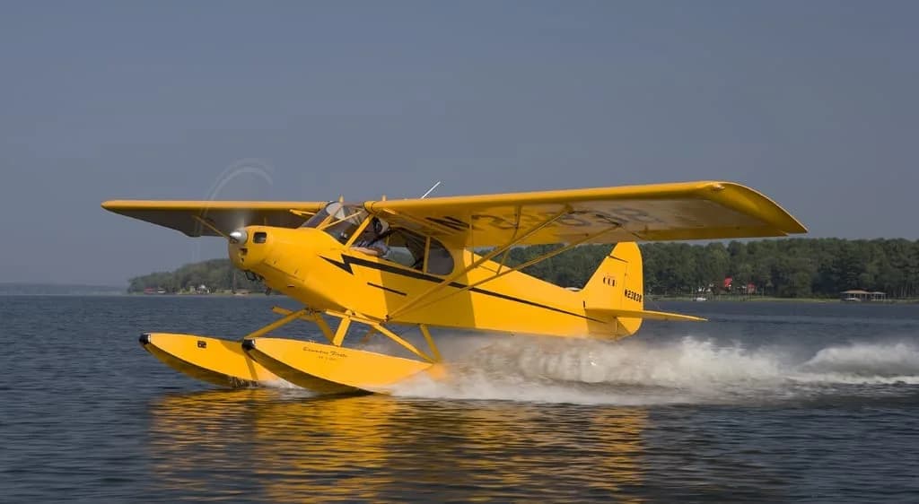 A yellow floatplane (N23838) with black stripes creates a wake as it moves across the water.